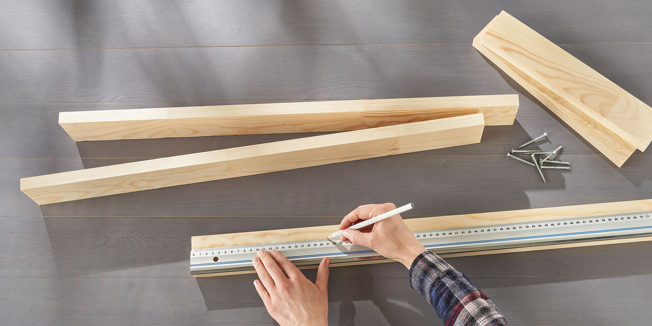 Hände zeichnen mit Bleistift und Lineal eine Linie auf ein Holzbrett. Auf dem Tisch liegen weitere Bretter und Schrauben.