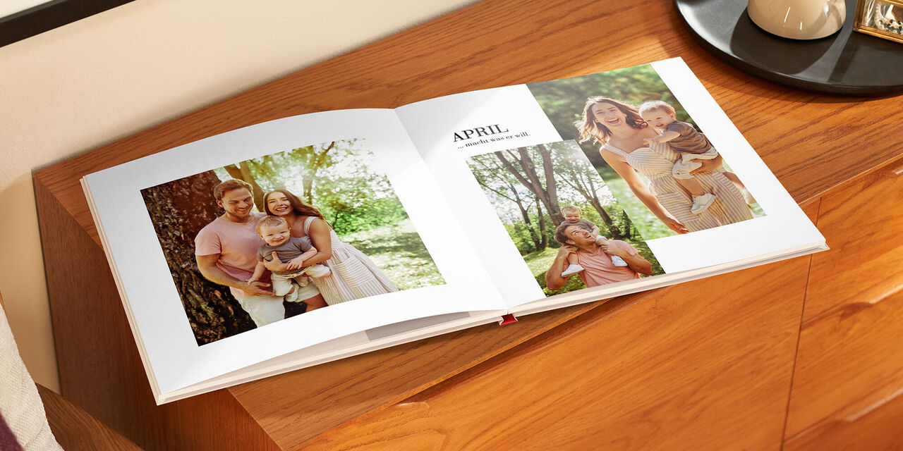Auf einem Holzschrank liegt das aufgeklappte Fotobuch. Auf der linken Seite ist ein Familienporträt mit Vater, Mutter und Kind zu sehen. Auf der rechten Seite befinden sich zwei Bilder, auf denen jeweils Mutter und Vater mit dem Kind abgebildet sind. Darüber steht die Überschrift «April».